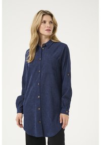 Chemise à manches longues bleu marine avec un col boutonné, une poche avant et un ourlet arrondi. Présente des boutons dorés et un tissu texturé.