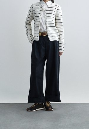 Woman wearing white patterned cardigan over white buttoned shirt, black wide-leg pants with brown belt, and dark sneakers standing on gray floor.