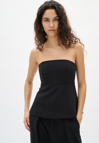Frau mit lockigem Haar, die ein trägerloses schwarzes Top und schwarze Hosen trägt, steht mit einer Hand in der Tasche vor einem schlichten weißen Hintergrund.