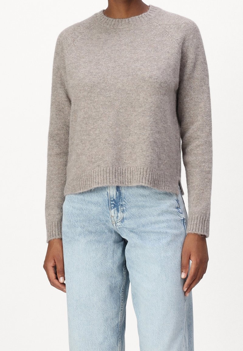 Personne portant un pull en maille à manches longues gris clair et un jean taille haute bleu clair, debout devant un fond uni.