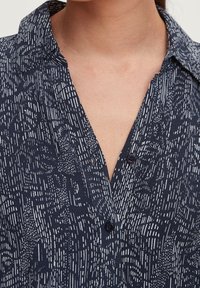 Mörkblå skjorta med knappar och en subtil textur i vitt. Har en krage, v-ringning och mörka knappar på knapplisten.