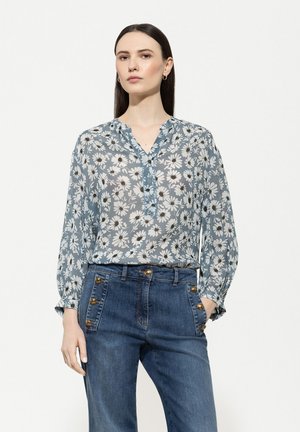 Hellbluse mit floralem Muster, langen Ärmeln, einem V-Ausschnitt und Knopfdetails, kombiniert mit hochtaillierten Jeans aus Denim mit goldenen Designelementen.