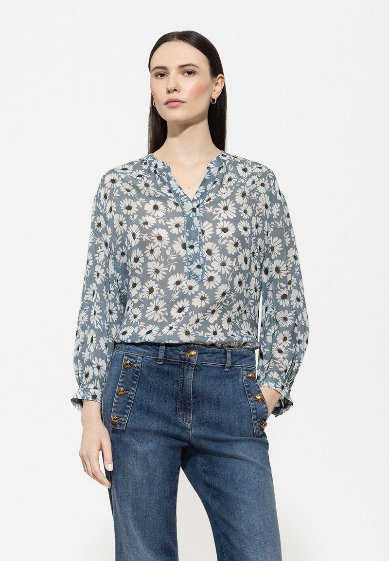 Hellbluse mit floralem Muster, langen Ärmeln, einem V-Ausschnitt und Knopfdetails, kombiniert mit hochtaillierten Jeans aus Denim mit goldenen Designelementen.