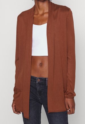 Žena v otevřeném hnědém kardiganu přes bílý krátký top a tmavě modré džíny, stojící před jednoduchým světlým pozadím.