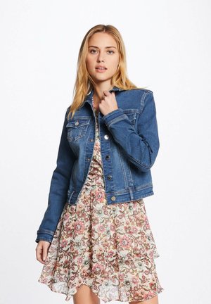 Veste en jean de bleu moyen, avec un col classique, des poches avant, une fermeture à boutons et des détails aux poignets, portée sur une robe à fleurs.
