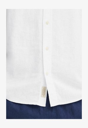 Bela lanena srajca z zakopčanjem na gumbe, s svetlobarvnimi gumbi in etiketo na spodnji levi strani. Nosena čez temno modre hlače.