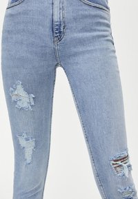 Vaqueros skinny de mezclilla azul claro con detalles desgastados y parches deshilachados. Diseño de cintura alta, estilo tradicional de cinco bolsillos y costuras visibles.