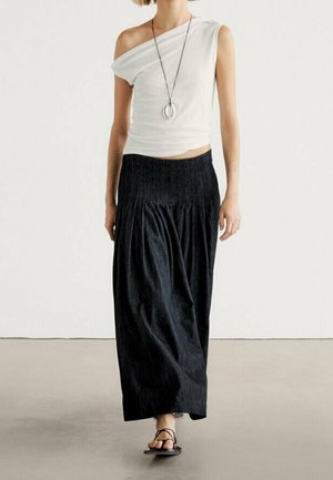 Femme portant un haut blanc asymétrique, une longue jupe noire plissée, des sandales noires à lanières et un long collier avec des pendentifs circulaires.