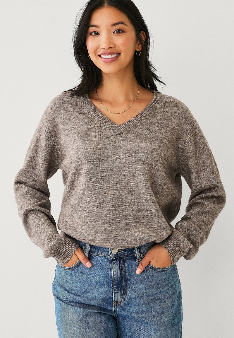 Lächelnde Frau mit langen dunklen Haaren trägt einen grauen V-Ausschnitt-Pullover und blaue Jeans, die Hände in den Taschen steckend, vor einem schlichten Hintergrund stehend.