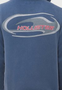 Μπλε σκούρο φούτερ με κουκούλα από βαμβάκι, με μεγάλο γκρι κυκλικό γραφικό και το "HOLLISTER" με κόκκινα γράμματα. Μαλακή υφή και κανονική εφαρμογή.