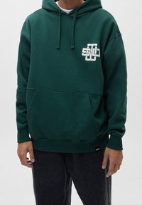 Sudadera de color verde oscuro hecha de una tela suave, con un bolsillo frontal, capucha ajustable y un detalle de logo blanco en el lado izquierdo del pecho.
