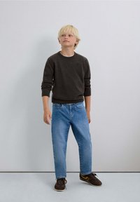 Pull col rond gris foncé, jeans droit bleu clair, chaussures marron avec des accents jaunes ; garçon se tenant contre un fond neutre.