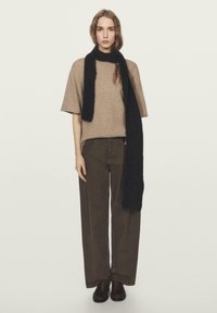 Kobieta o falowanych włosach ubrana w beżowy sweter z krótkim rękawem, ciemnobrązowe spodnie o szerokich nogawkach, czarny szalik i ciemnobrązowe buty na jednolitym tle.