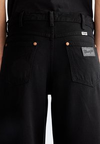 Vue arrière d'un jean Wrangler noir avec deux poches arrière, des rivets en cuivre, des passants pour ceinture et une étiquette Wrangler sur la poche droite.