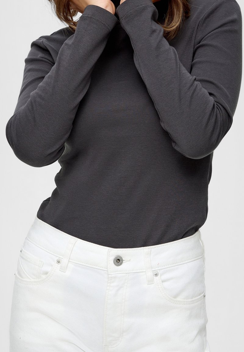 Haut en coton à manches longues, gris foncé, avec un col haut, porté avec un jean en denim blanc présentant une fermeture à bouton standard et des poches.