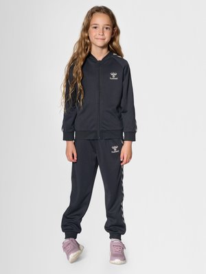 Jong meisje met lang haar staat in een zwart Hummel trainingspak en roze sneakers op een effen lichte achtergrond.