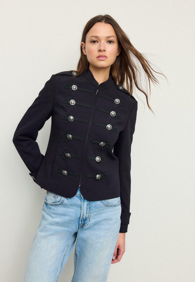 Femme aux longs cheveux bruns portant une veste militaire noire avec des boutons argentés et un jean bleu clair, debout devant un fond uni.