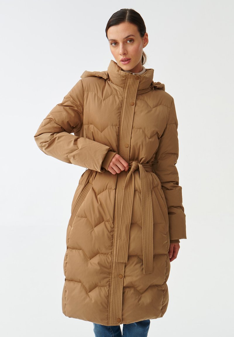 Brązowy, pikowany płaszcz z wysokim kołnierzem, paskiem w talii i zapięciem na guziki. Posiada szycia w zygzak oraz prążkowane detale z przodu.