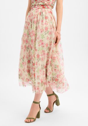 Femme portant une jupe midi fluide et transparente à motifs floraux roses et verts, associée à des talons blocs à bride cheville couleur vert olive.