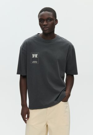 Junger Mann mit kurzen Haaren, der ein dunkelgraues, übergroßes T-Shirt mit kleinem weißem Logo und beige Hosen trägt, vor einem schlichten Hintergrund stehend.