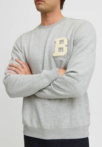 Sudadera gris hecha de una tela suave, con un emblema texturizado de color crema en forma de "B" en el pecho, escote redondo y puños acanalados.