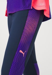 Gros plan d’une jambe portant des leggings bleu marine avec des panneaux latéraux à motifs violets et roses comportant un logo Puma rose et un top assorti violet et rose.