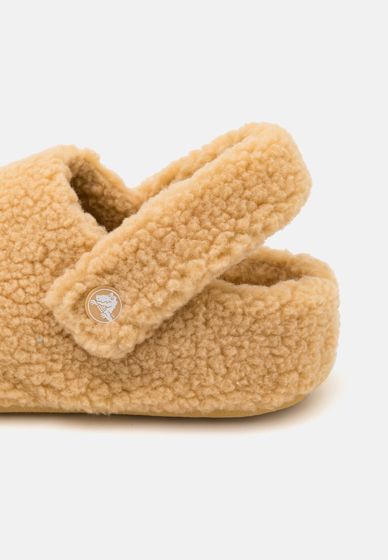 Chausson en polaire beige avec une surface texturée, présentant une large sangle et un accent de logo circulaire. Design doux et confortable avec une forme arrondie.