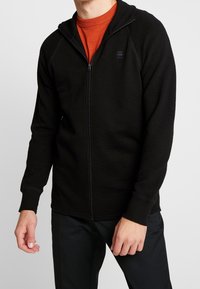 Sudadera con cremallera negra que presenta una tela texturizada, acentos acanalados y un diseño ajustado. Combinada con una camiseta naranja de cuello redondo debajo.