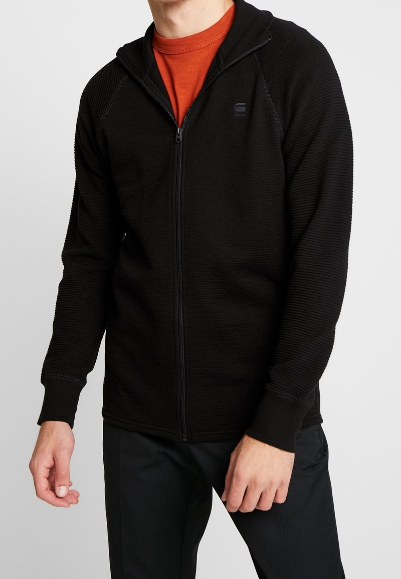 Sudadera con cremallera negra que presenta una tela texturizada, acentos acanalados y un diseño ajustado. Combinada con una camiseta naranja de cuello redondo debajo.