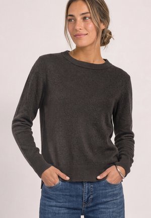 Femme aux cheveux châtain clair en chignon, portant un pull en maille gris foncé et un jean bleu, mains dans les poches, regardant sur le côté et souriant légèrement.