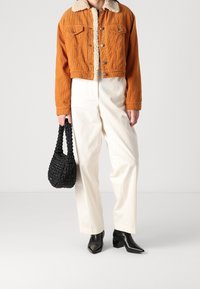 Giacca corta in velluto a coste arancione con fodera in pile, abbinata a pantaloni bianchi a gamba larga e borsa nera intrecciata. Stivaletti neri con punta affilata.