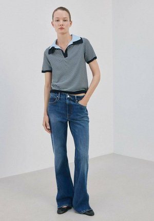 Joven de pie vestida con una camisa tipo polo de rayas blancas y negras, jeans azules de corte acampanado y zapatos negros puntiagudos en una habitación sencilla.