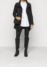 Svart texturerad kappa med slag, två knappar, bärs över en vit skjorta. Svarta läderleggings och knähöga stövlar fulländar looken.