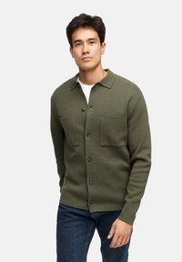 Olivgrön texturerad cardigan med knapstängning, två bröstfickor och krage, ihopparad med mörka jeans.