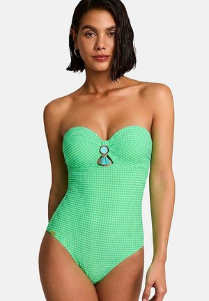 Femme aux cheveux courts et foncés portant un maillot de bain vert texturé sans bretelles avec un anneau décoratif et un pompon au niveau de la poitrine.