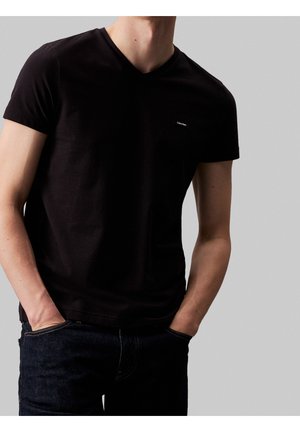 Personne portant un T-shirt noir uni à col en V et manches courtes, avec un petit logo sur la poitrine, mains dans les poches d’un jean bleu foncé.