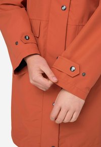 Vesitiivis oranzhi takki, jossa on suuret kuusikulmaiset napit hihansuissa, etutasku ja sileä pinta.
