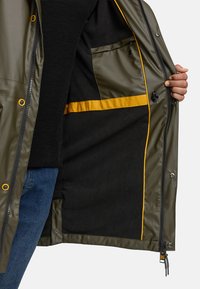 Waterdichte olijfgroene jas met ritssluiting, gele accenten, een gladde buitenkant en een zachte zwarte binnenvoering met zakken.