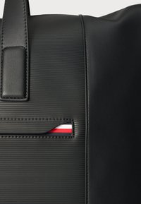 Geantă neagră cu suprafață texturată și panouri din piele netedă. Include un buzunar mic care afișează un card cu accente roșii și albastre.