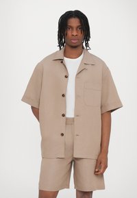 Beige Kurzarmhemd mit fünf braunen Knöpfen, einer Brusttasche und passenden Shorts. Glatte Textur, lässiges Design. Weißes Unterhemd sichtbar.