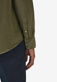 Olivi vihreä pitkähihainen paita, jossa napitettavat hihansuut, valmistettu sileästä kankaasta. Pyöreä helma näkyvissä, yhdistettynä tumman laivastonsiniseen alaosaan.