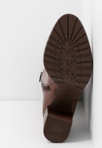 Bottine en cuir marron avec une semelle en caoutchouc texturée, dotée d'une fermeture éclair et d'un talon bloc, présentée de côté sur une surface blanche.
