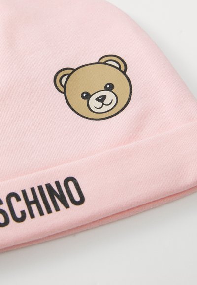 Světle růžová textilní čepice s usmívající se tváří mé teddy bear a částečně viditelným černým textem "MOSCHINO" na ohnutém okraji.