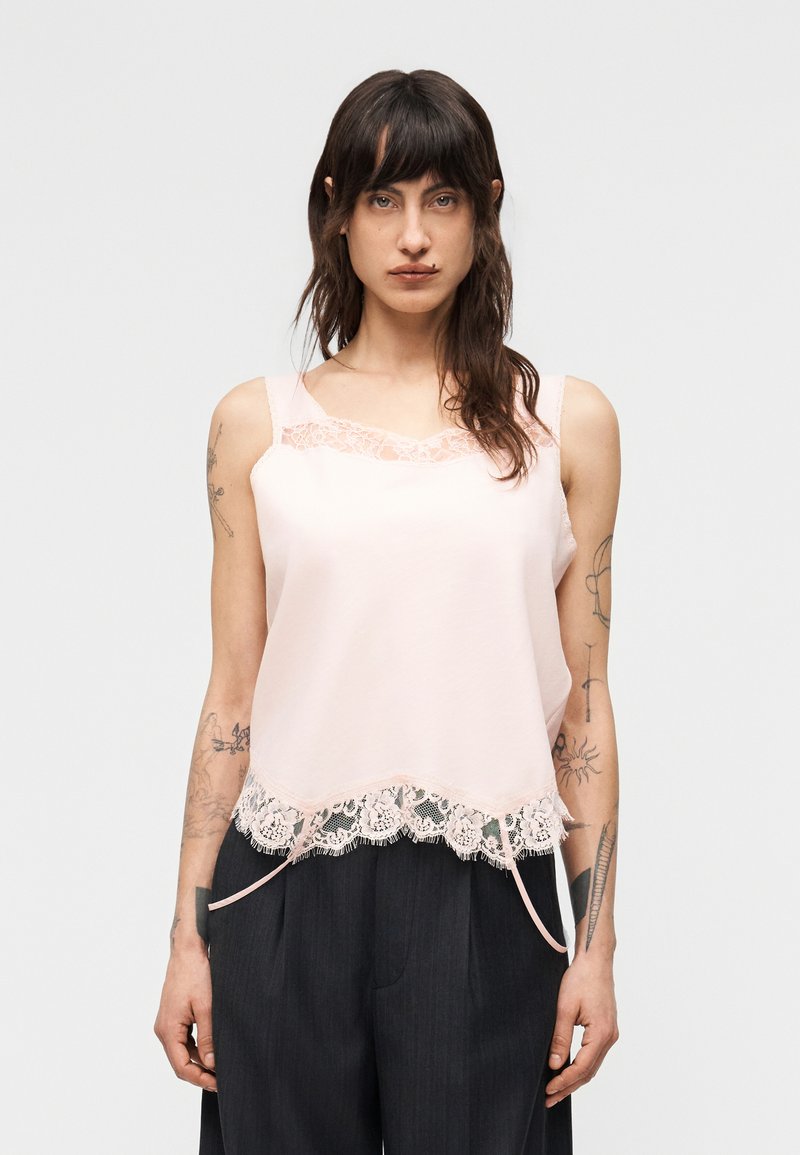 Γυναίκα με σκούρα μαλλιά και τατου άζ φοράει απαλό ροζ αμάνικο τοπ με δαντέλα και σκούρο φαρδύ παντελόνι μπροστά σε απλό φόντο.