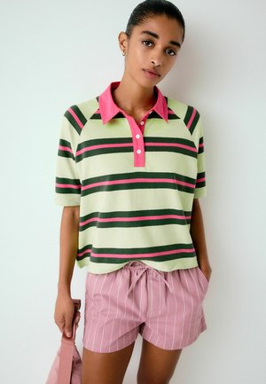 Junge Frau trägt ein grünes und pink gestreiftes Poloshirt und pink gestreifte Shorts, hält eine passende pinke Tasche an ihrer Seite.