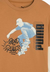 Braunes Baumwoll-T-Shirt mit einem Grafikdesign eines Skateboardfahrers in Blau, mit dem Text "BEND THE RULES" und "PUMA" in Schwarz.