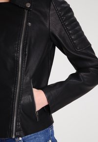 Svart konstläderjacka med vadderade axlar, silverfärgad framdragkedja och sidofickor med dragkedja, som bärs med blå denimjeans.