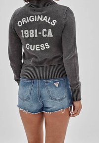 Szary sweter z dzianiny z prążkowanym kołnierzem, z białym napisem „ORIGINALS 1981-CA” i „GUESS” na plecach, zestawiony z niebieskimi dżinsowymi szortami z przetarciami.