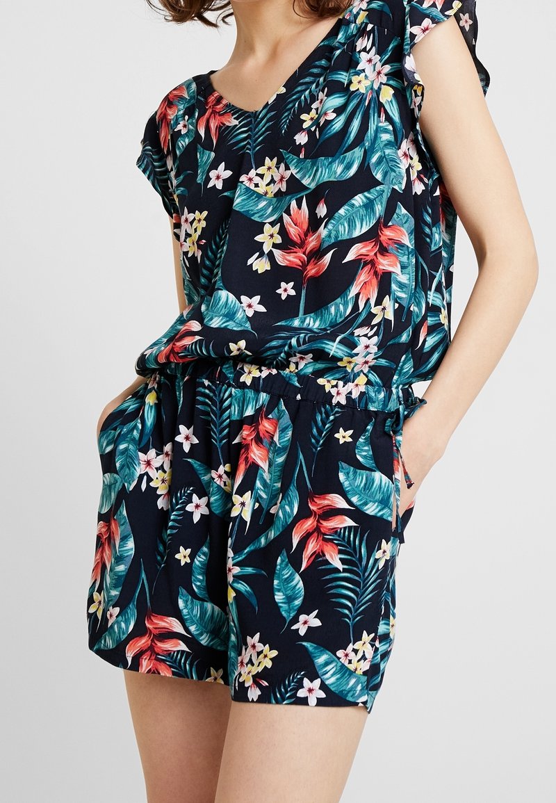 Femme portant une combishort à manches courtes bleu marine avec des motifs floraux tropicaux rouges, blancs et verts, mains dans les poches.