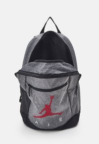 Pilkas audinys kuprinė su juodu pagrindu, turinti priekinį užtrauktuką, tinklines šonines kišenes ir raudoną Jumpman logotipą su "AIR" tekstu.